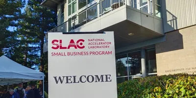 A welcome sign for the SLAC National Accelerator Laboratory Small Business Program, set against the backdrop of a modern building and lush trees, with a white tent and people gathered in the foregroun