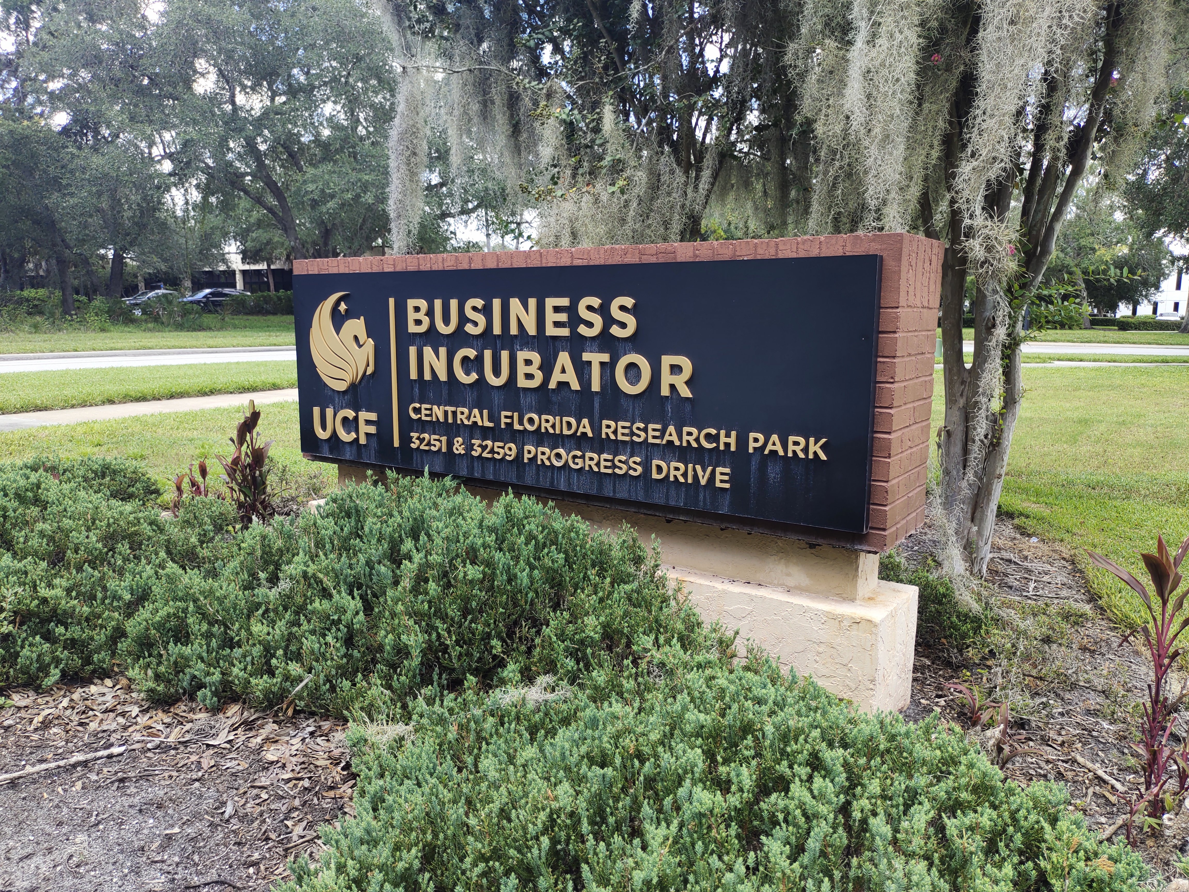 A close-up shot of the UCF Business Incubator sign located at Central Florida Research Park. The brick-framed sign features gold lettering on a black background amidst green shrubbery and hanging Spanish moss.