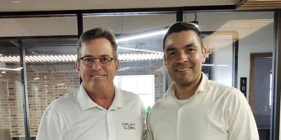 Two business professionals stand side-by-side in a modern office environment. The man on the left wears a white polo shirt with the Gardant Global logo, while the man on the right wears a light-colored button-down shirt. They are smiling in front of a glass-walled conference room with warm brick accents.