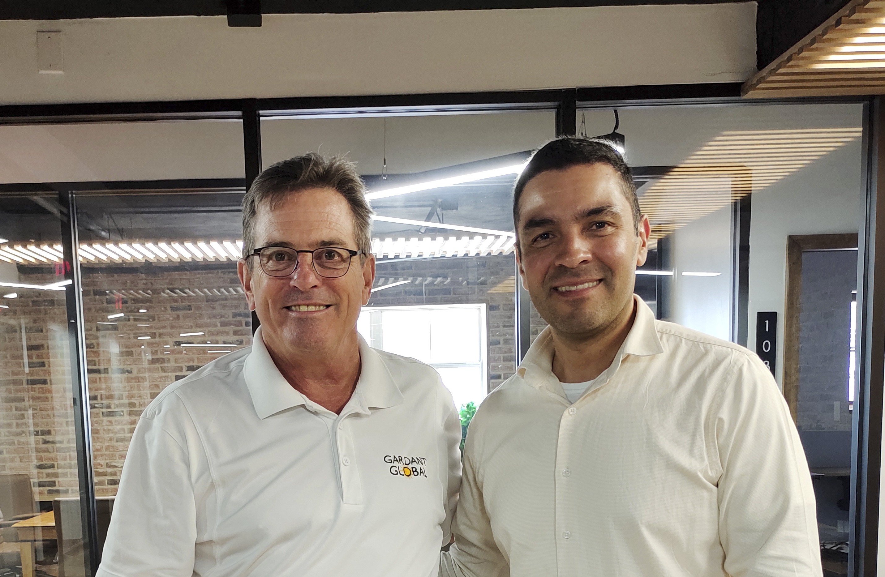 Two business professionals stand side-by-side in a modern office environment. The man on the left wears a white polo shirt with the Gardant Global logo, while the man on the right wears a light-colored button-down shirt. They are smiling in front of a glass-walled conference room with warm brick accents.