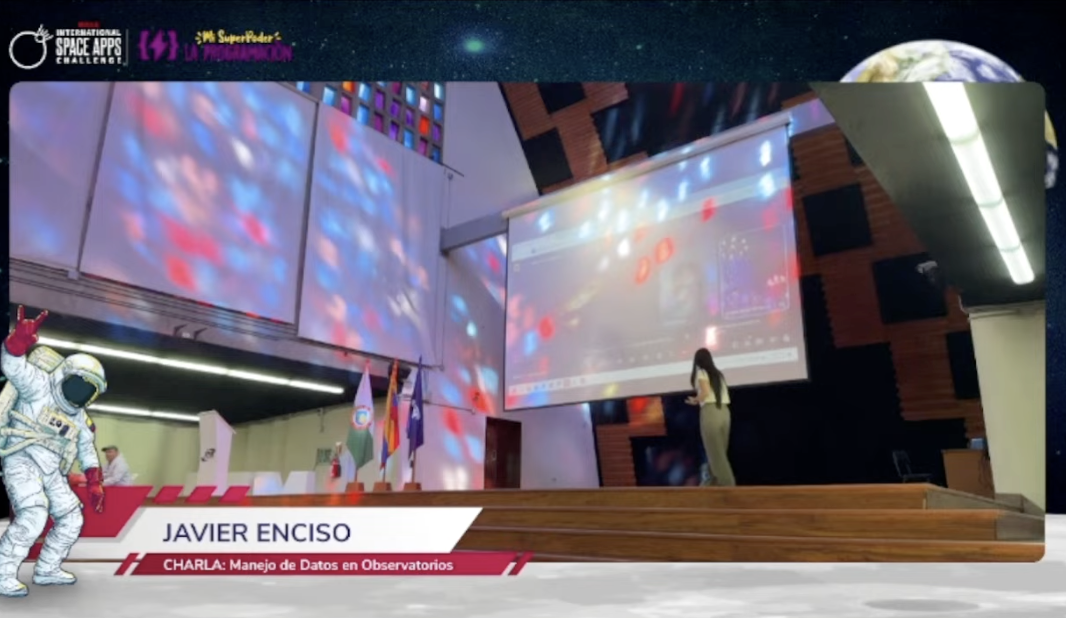 A speaker, Javier Enciso, presents on 'Data Management in Observatories' at the 2023 International Space Apps Challenge. The stage includes a projector screen displaying web content, flags, and a playful astronaut graphic, all set against a space-themed b