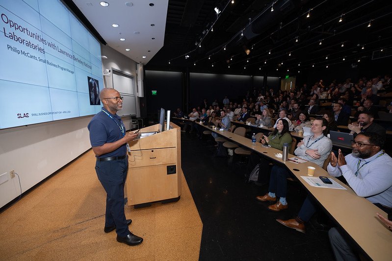 Philip McCants, Small Business Program Manager, presents on opportunities with Lawrence Berkeley National Laboratories at the SLAC Small Business Opportunity conference. He addresses an engaged audience in an auditorium, many of whom are applauding.